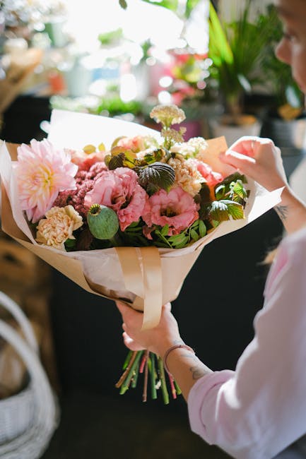 A person holding a large, fresh floral bouquet wrapped in beige paper with a cream ribbon. The bouquet features a mix of pink and white flowers, including peonies, carnations, and chrysanthemums, acce