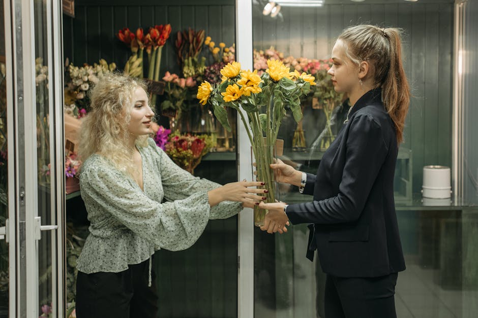 A customer receives a bouquet arrangement of bright yellow sunflowers and smaller pink flowers from a florist at a flower shop, with the florist dressed in a dark blazer and the customer in a patterne