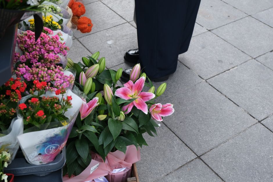 A close-up view of a floral arrangement featuring pink Asiatic lilies with partially open petals and prominent dark stamens, surrounded by lush green leaves. The lilies are placed among other fresh fl