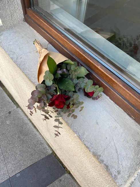 A small bouquet with two red roses, eucalyptus leaves, and other green foliage, wrapped in light brown paper, rests on a concrete ledge beneath a window with a wooden frame. The bouquet appears freshl