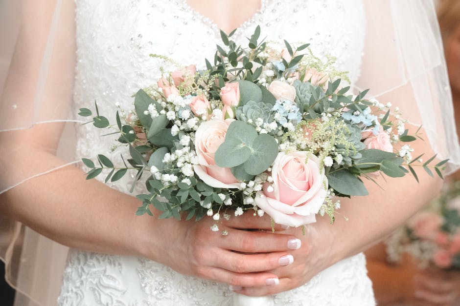 A bride is holding a floral bouquet arrangement featuring large, soft pink roses, small white baby's breath, and eucalyptus leaves, with a variety of greenery adding texture. The bouquet is wrapped in