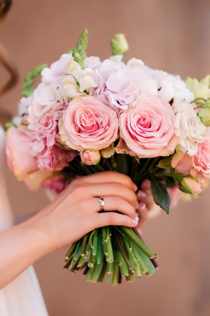 A close-up of a bride's hand holding a bouquet of fresh flowers, featuring soft pink roses, white lisianthus, and green foliage, arranged in a rounded style. The bouquet is wrapped with a simple green